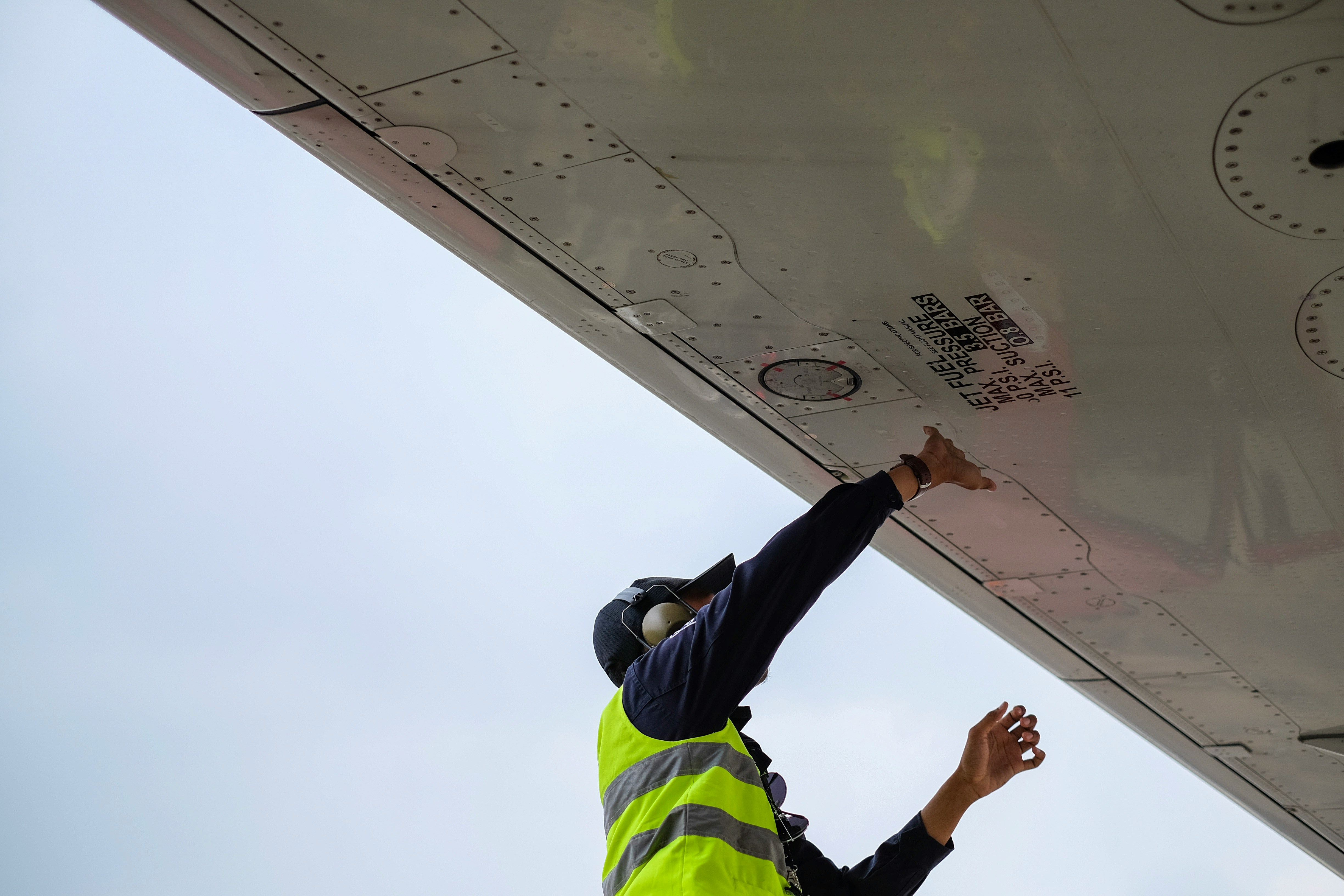 A&P apprentice mechanic in hi-vis vest inspecting an aircraft wing fuel panel on the flight line